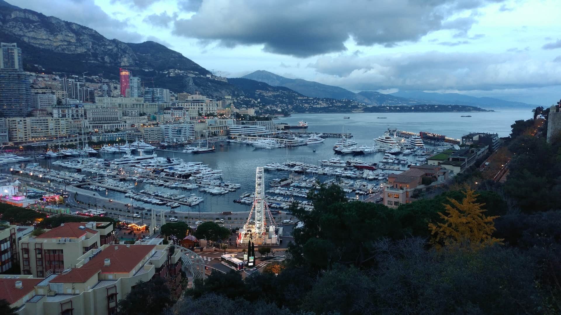 Зимний вечер в монекасской гавани с роскошными яхтами, подсвеченным колесом обозрения и драматичными облаками над горами.
