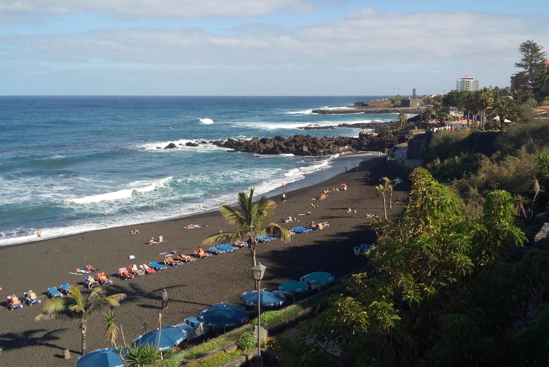 Солнечный пляж с чёрным песком на Тенерифе в январе, с отдыхающими на шезлонгах, купающимися в Атлантике людьми и накатывающимися на берег волнами.
