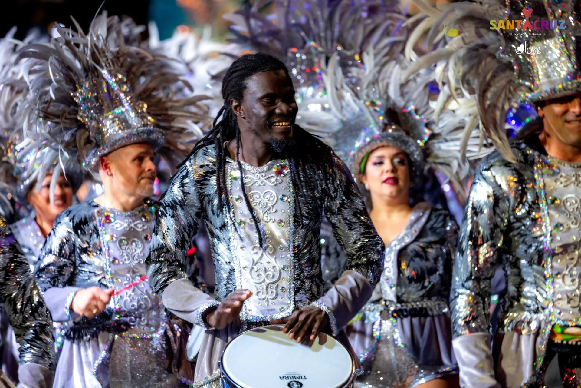 A group of Carnival performers in shiny silver costumes and massive feather headpieces; in the foreground, a smiling man with dreadlocks plays a drum during a festive parade.