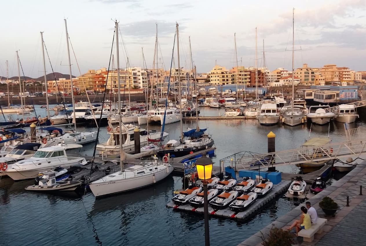 Fotografía paisajística elevada de Marina del Sur en Las Galletas, Tenerife, que muestra un puerto dinámico lleno de numerosos veleros y yates a motor blancos en los pantalanes, respaldado por un pueblo de edificios de baja altura y colinas suaves bajo la luz difusa del atardecer. En primer plano hay motos de agua alineadas, una farola encendida y una pareja relajada en el banco del paseo marítimo.